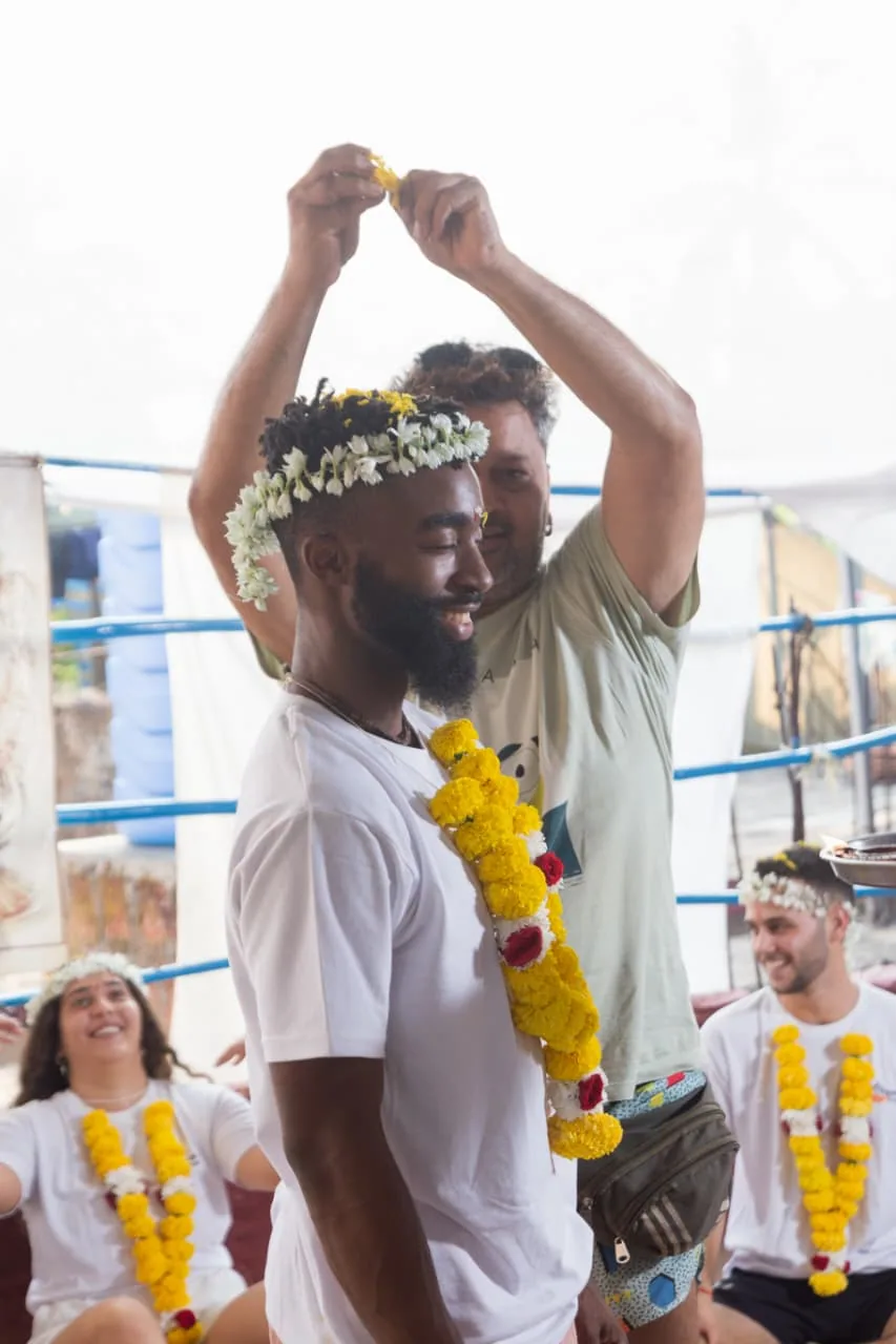 Graduate receiving a jasmine flower crown from his teacher while garlanded classmates laugh behind him