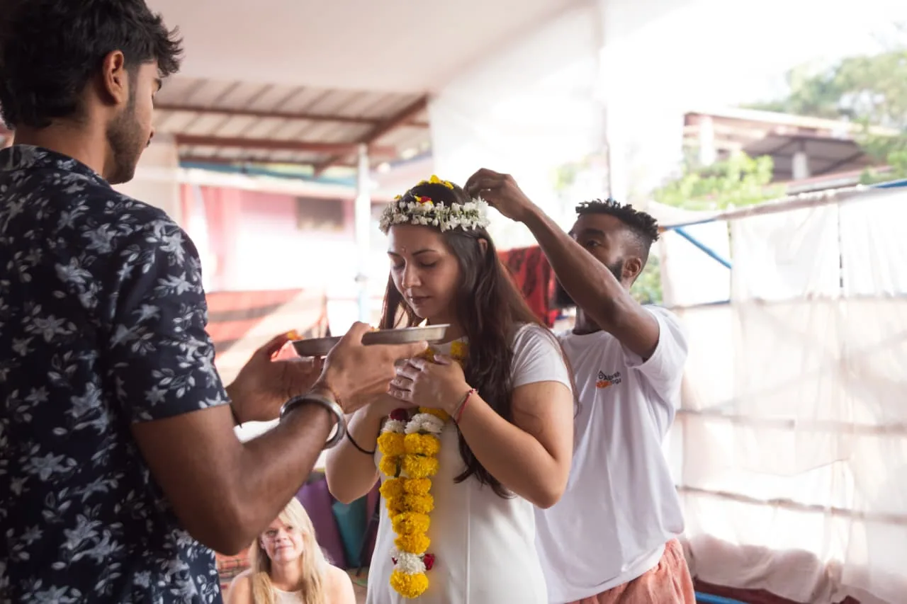 Yoga teacher training graduate receiving a jasmine flower crown and marigold garland while a teacher holds the puja plate at the closing ceremony