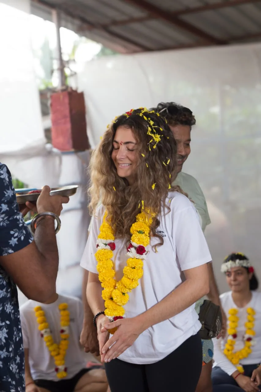 Smiling graduate wearing a marigold garland, flower petals in her hair, receiving a blessing at the closing ceremony of her yoga teacher training