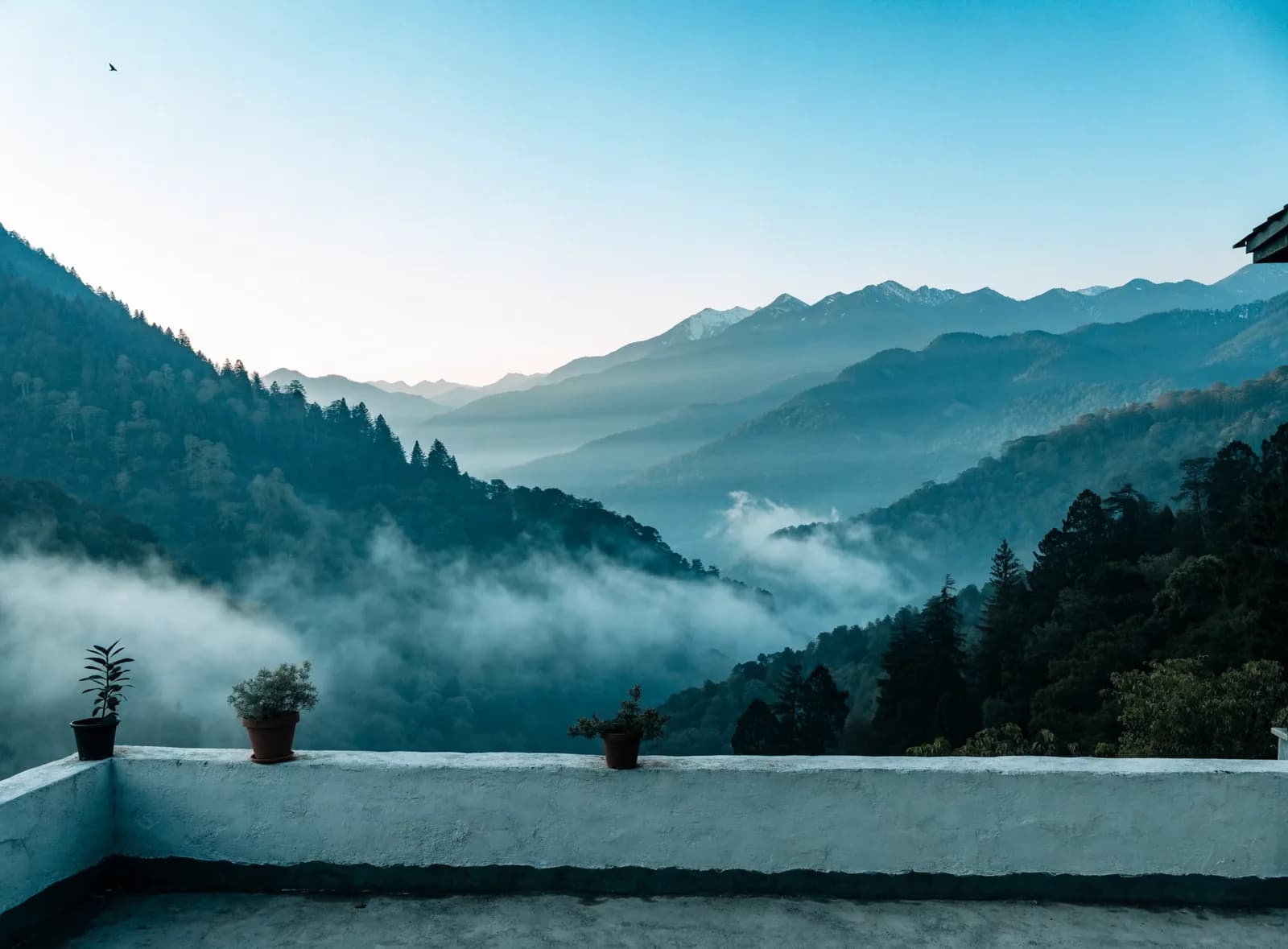 The view from a flat rooftop terrace over a Himalayan valley at dawn, mist between the forested ridges and snow on the far peaks