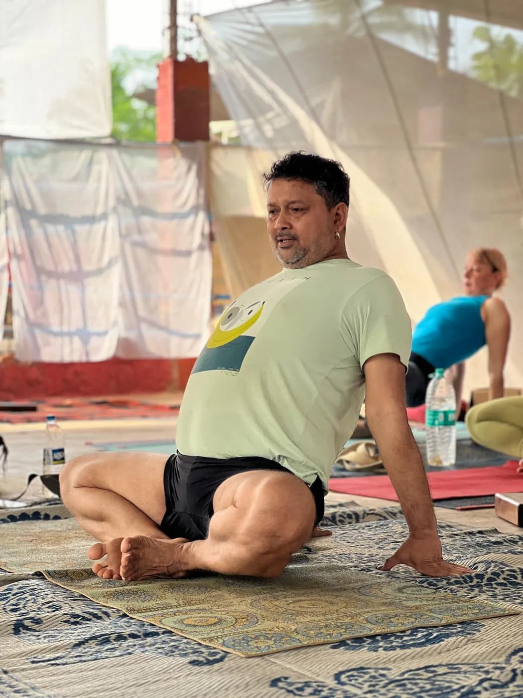 Alpesh Patwari seated on a mat talking through a seated twist while students practise behind him
