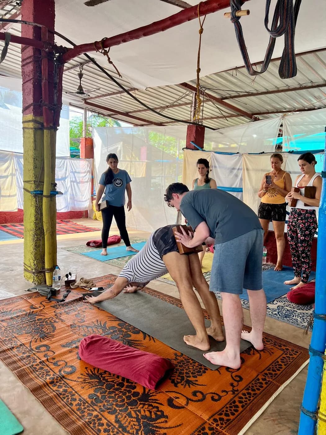 Hands-on assist in a standing forward fold using a wooden block, watched by the rest of the class during teaching practice at the shala