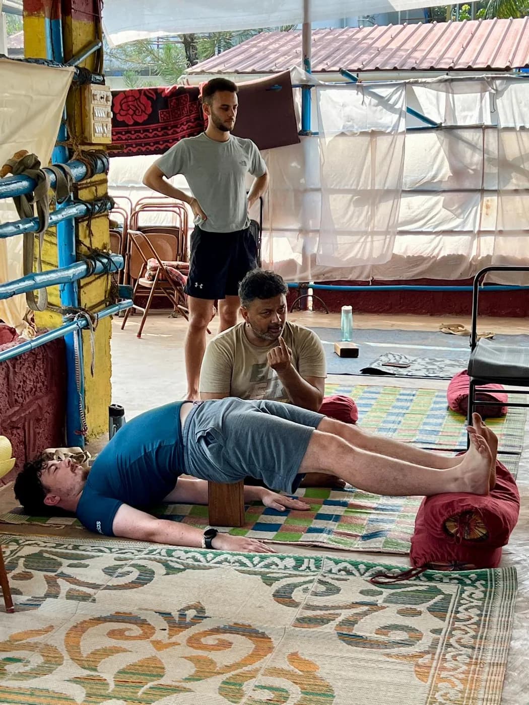 Student in a supported bridge pose with a wooden block under the sacrum and a bolster under the heels while a teacher explains the alignment